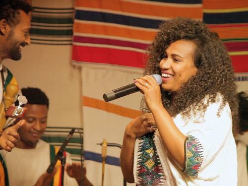 eine junge Frau singt. Sie hält ein Mikrophon in der Hand. Am linken Bildrand sieht man zwei Musiker.